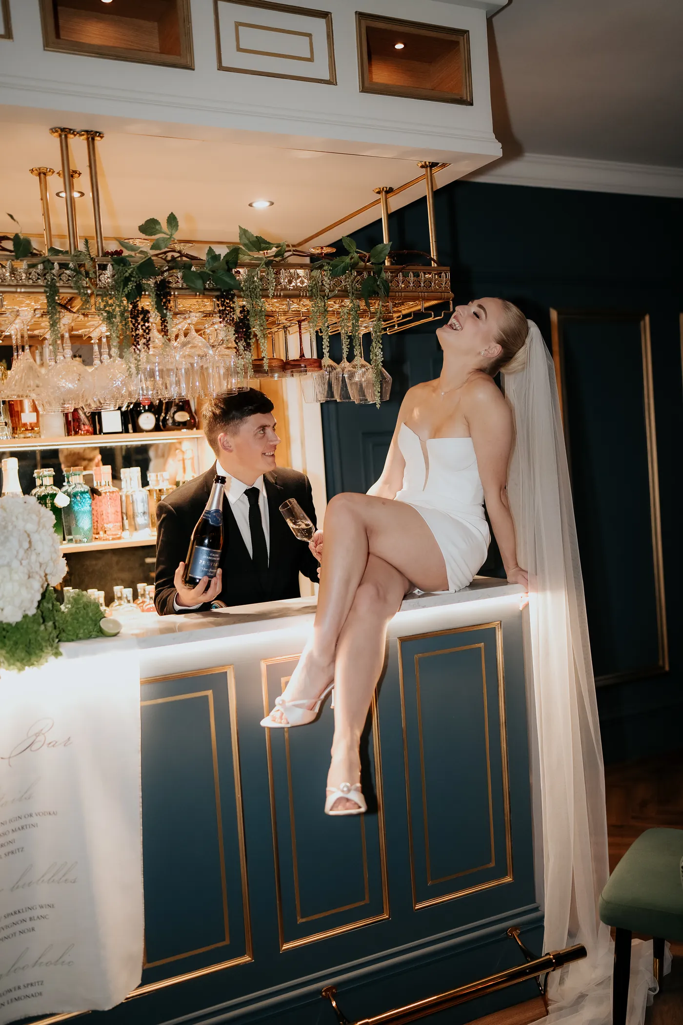 A bride sitting on the bar in Nocturne, laughing, while the bartender opens a bottle