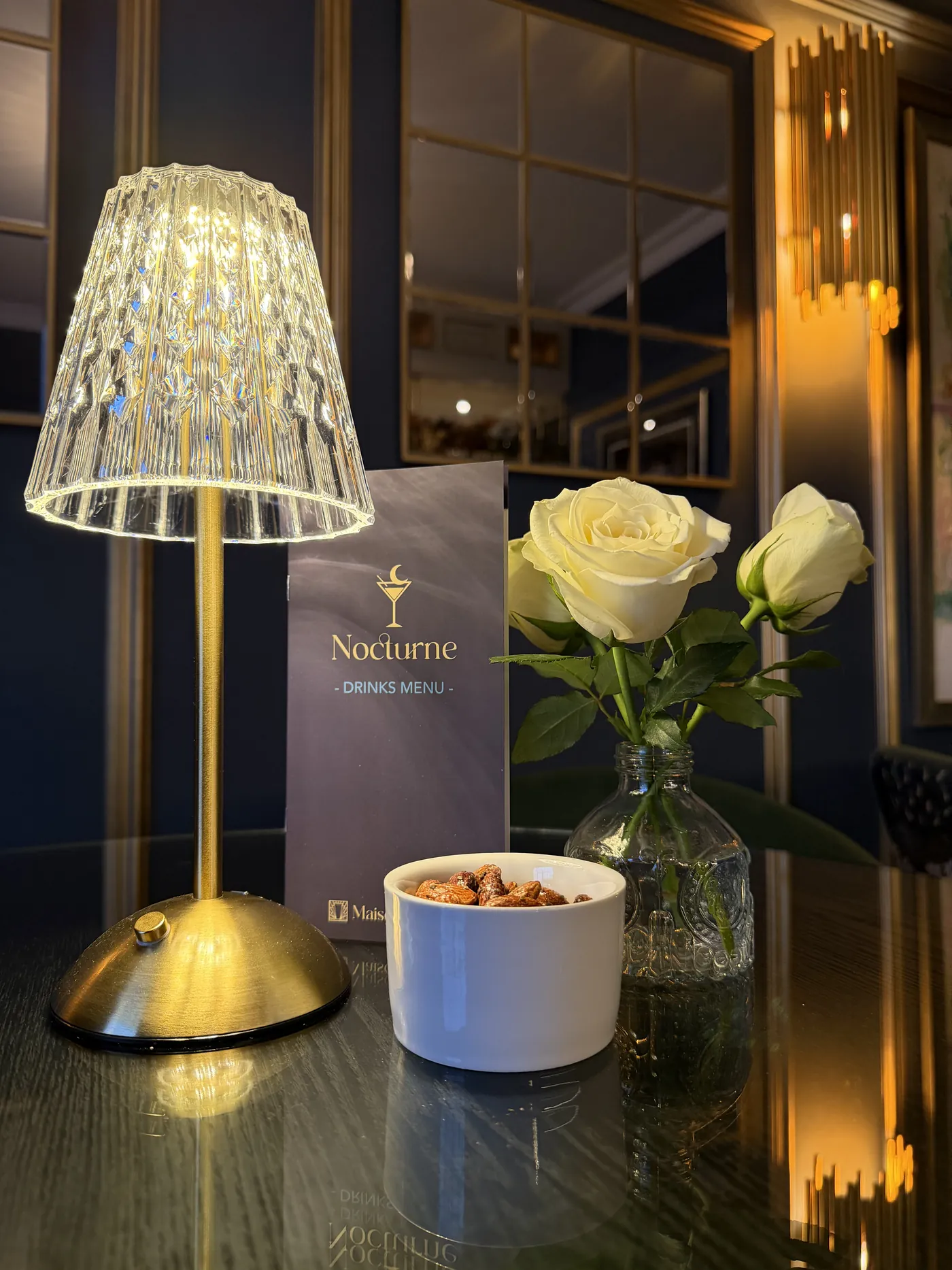 A crystal table lamp, a white rose and the Nocturne drinks list on a black glass table