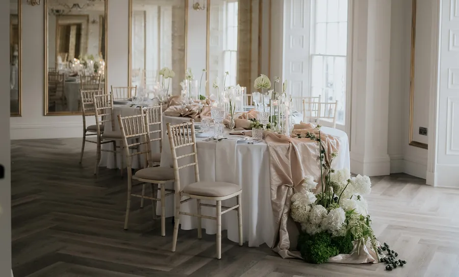 Orchidée dressed for a celebration dinner with blush linen, gold chairs and white flowers by tall windows