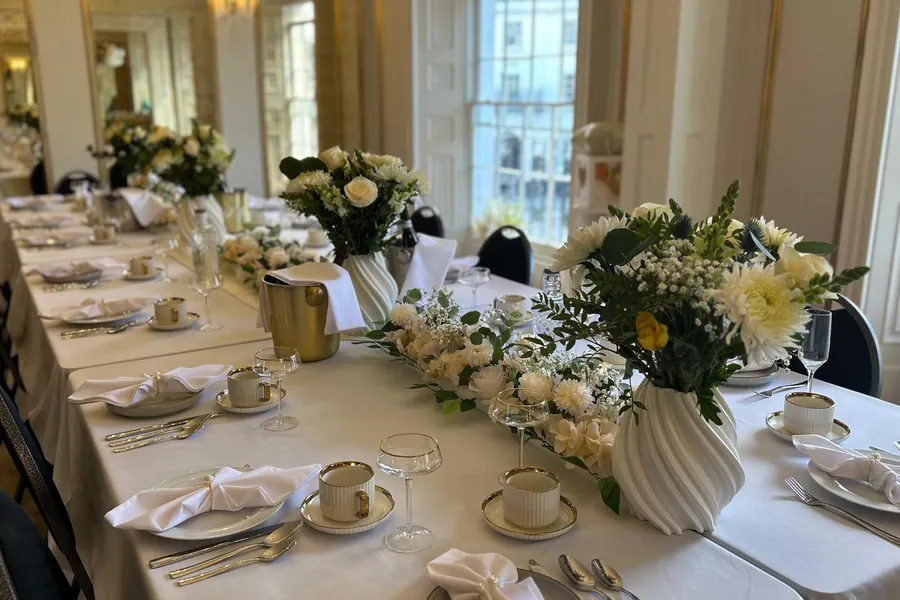 A long table laid with white linen, gold cutlery, cups and white flowers by a tall window